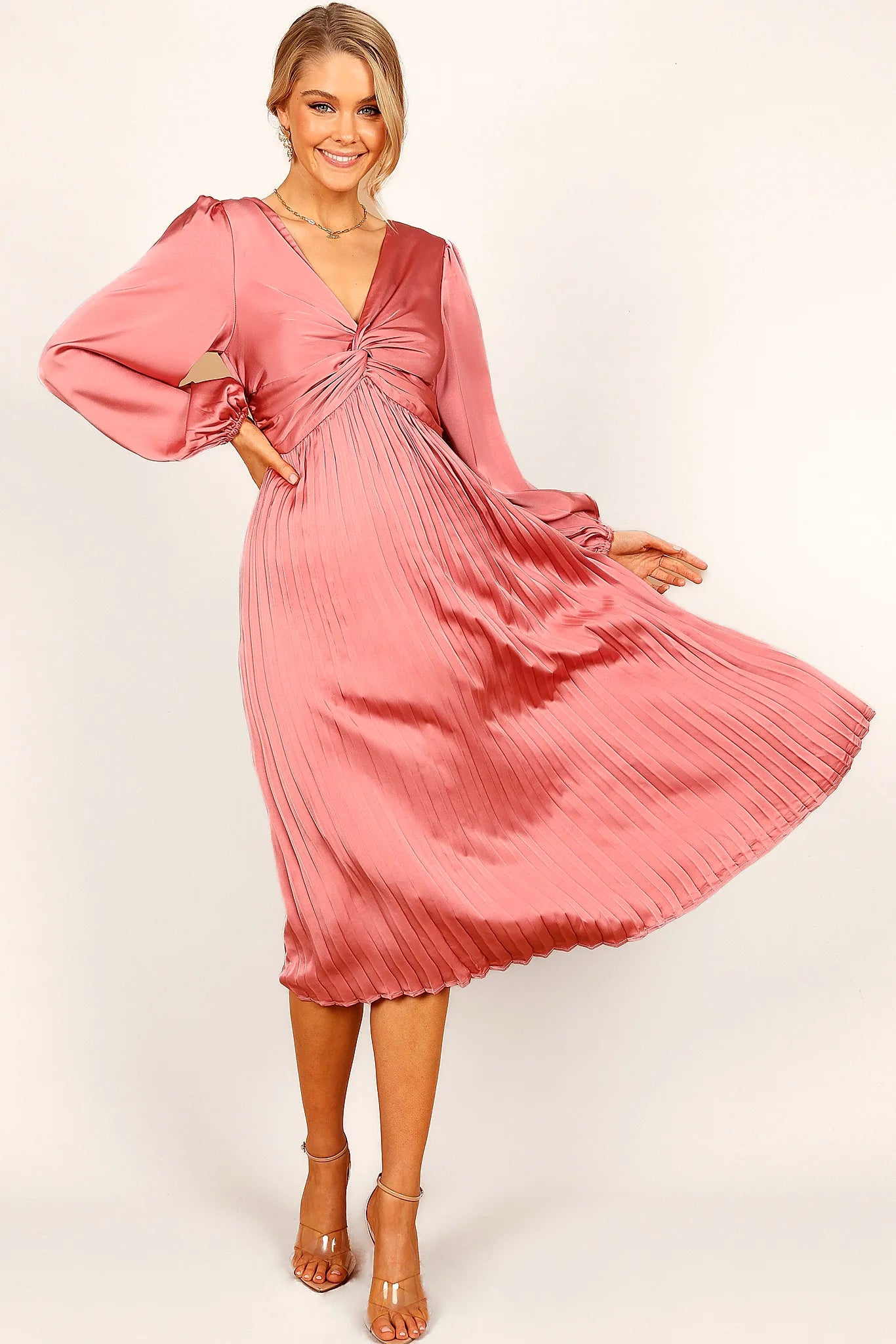 Side view of a pink blush twist front pleated midi dress, worn by a model against a plain studio backdrop