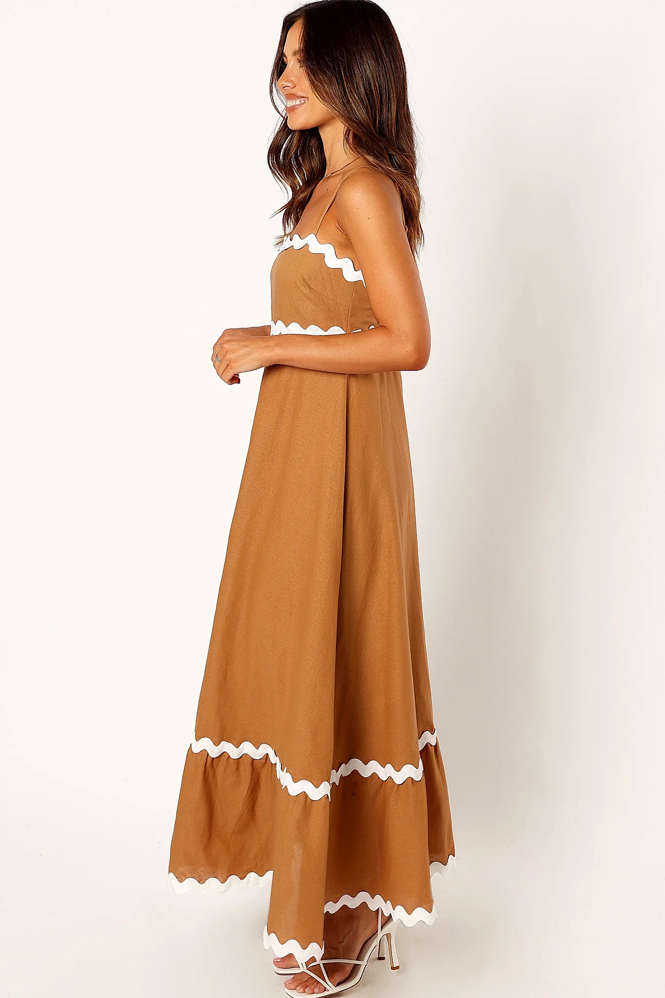 Side view of a white tan & white maxi dress, thin straps, worn by a model against a plain studio backdrop